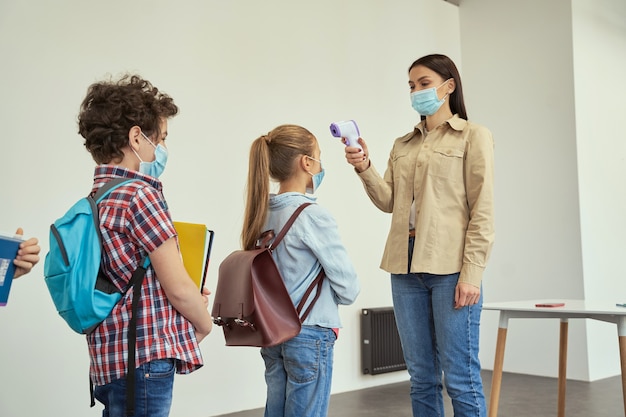 Foto professora usando máscara protetora, examinando crianças em idade escolar em busca de febre contra a propagação de