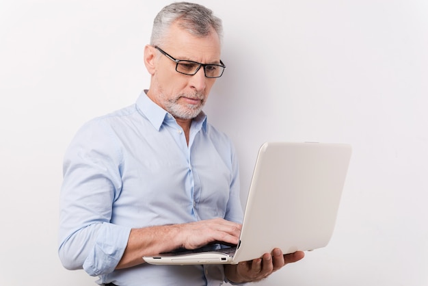 Profi bei der Arbeit. Selbstbewusster älterer Mann in Hemd und Brille, der am Laptop arbeitet, während er vor grauem Hintergrund steht