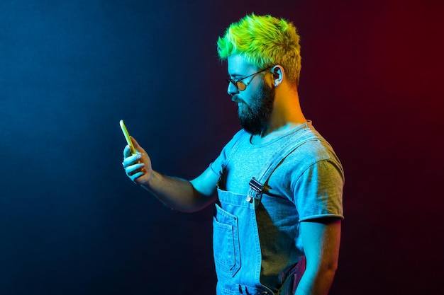 Profil eines jungen, bärtigen Hipster-Typen mit grünen Haaren, der Denim-Overalls trägt, Smartphone in den Händen hält, blickt mit ernster Miene auf die Anzeige. Buntes Neonlicht, Studioaufnahme im Innenbereich.