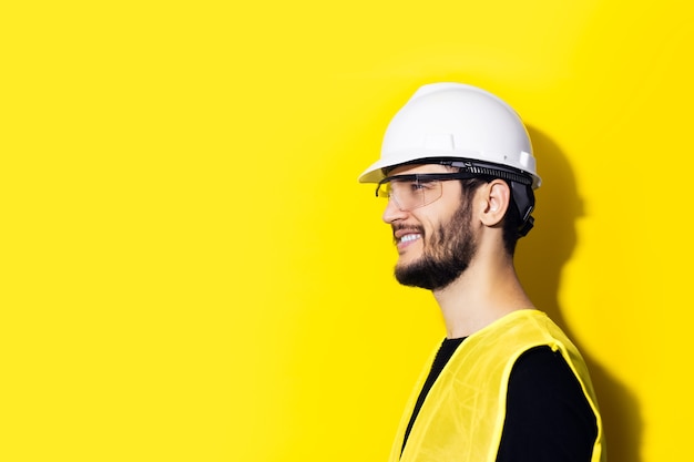 Profil eines jungen lächelnden Mannarchitekten, Baumeisteringenieurs, tragend weißen Konstruktionssicherheitshelm, Brille und gelbe Jacke lokalisiert auf gelber Wand.