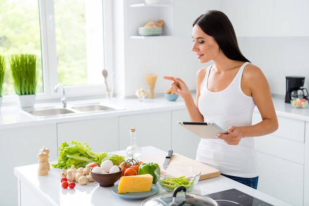 Profil Seite Foto von niedlichen süßen braunhaarigen Hausfrau wollen kochen Morgen Frühstück folgen Rezept Anzahl Zutaten Gemüse Käse Pilze rote Tomate verwenden Tablette in weißen Haus Küche