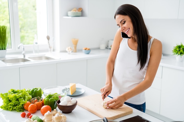 Profil Seite Foto von positiven fröhlichen Mädchen Hausfrau haben Ruhe entspannen wollen Mittagessen vorbereiten Gesundheitswesen Abendessen geschnitten Champignon auf hacken Holzbrett mit Freunden in Haus weiße Küche kommunizieren