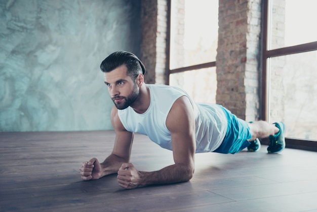 Profilfoto des Macho-Sportler-Mannes, der Planke auf Ellbogenhänden tut, die Boden lehnen, bestimmte Sportswear-Tanktop-Shorts Turnschuhe, die Hausfenster drinnen trainieren