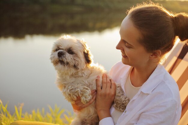 Profilporträt einer dunkelhaarigen Frau mit Brötchenfrisur, die ein weißes Hemd trägt, das mit ihrem Pekingese-Hund in der Nähe des Flusses sitzt und gemeinsam Spaß hat, wenn die Frau mit ihrem Haustier in der Natur spielt