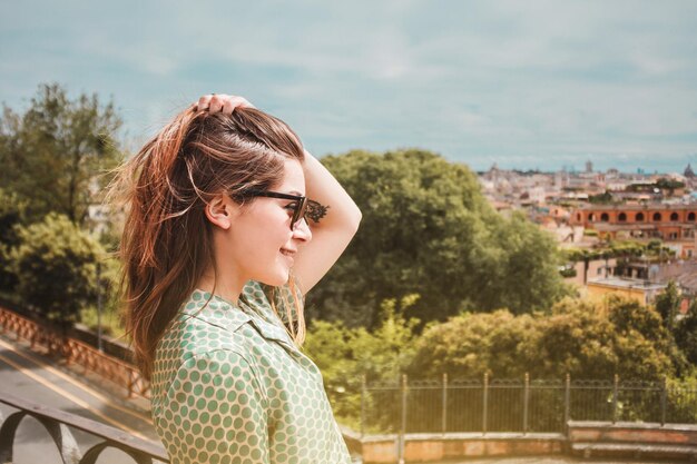 Foto profilporträt einer jungen lächelnden frau in sonnenbrille mit panoramablick auf rom, italien