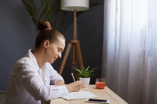 Profilporträt einer jungen schönen Frau, die ein weißes Hemd trägt und Notizen aufschreibt, während sie am Tisch vor dem Fenster sitzt. Feamle mit Brötchenfrisur schreibt im Organizer