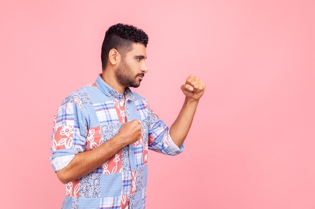 Profilporträt eines aggressiven jungen erwachsenen Mannes in blauem Hemd, der mit Boxfäusten steht und bereit zum Angriff oder zur Verteidigung ist und mit wütendem Gesicht aussieht Studioaufnahme im Innenbereich isoliert auf rosa Hintergrund