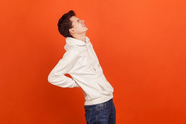 Profilporträt eines frustrierten, ängstlichen Teenagers in weißem Hoodie und lässigen Jeans, der die Hände am unteren Rücken hält, Nierenverletzung, Rückenschmerzen. Studioaufnahme im Innenbereich isoliert auf orangefarbenem Hintergrund