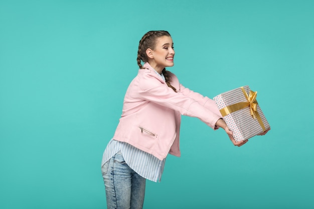 Profilseitenansicht eines glücklichen, schönen Mädchens im lässigen Stil, Zopffrisur und rosa Jacke, stehend und gepunktete Geschenkbox mit zahnigem Lächeln, Studioaufnahme einzeln auf blauem oder grünem Hintergrund