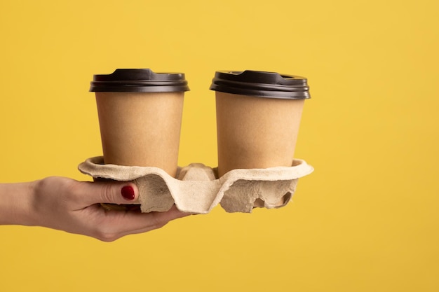 Profilseitenansicht, Nahaufnahme einer Frau, die eine Tasse heißes Bechergetränk zum Mitnehmen in der Hand hält und zeigt, Kaffeepause bei der Arbeit. Studioaufnahme im Innenbereich isoliert auf gelbem Hintergrund.