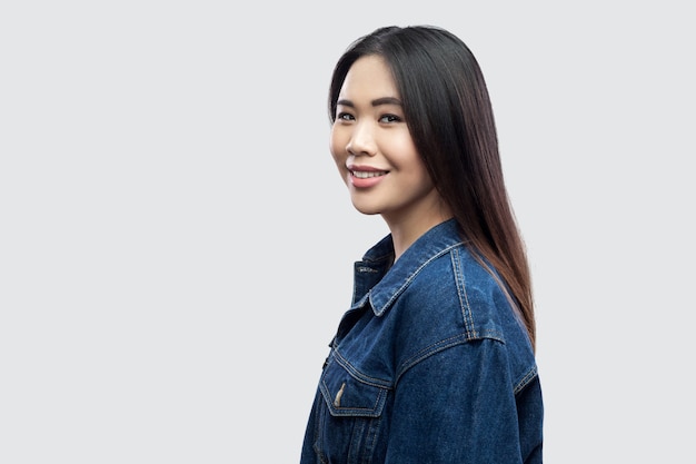 Profilseitenansicht-Porträt eines glücklichen, schönen, brünetten asiatischen Mädchens in lässiger blauer Jeansjacke mit Make-up-Zahnlächeln und Blick in die Kamera. Indoor-Studioaufnahme, isoliert auf hellgrauem Hintergrund.