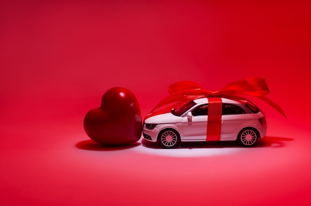 Profilseitennahaufnahmefoto eines schönen weißen Spielzeugs, modernes modisches trendiges Auto mit rotem Band und rotem Herz in der Mitte im Scheinwerferlicht einzeln auf rotem Hintergrundkopierraum