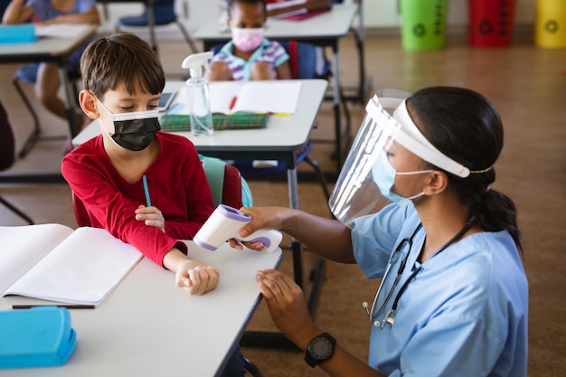 Profissional de saúde feminina usando protetor facial medindo a temperatura de um menino na escola primária