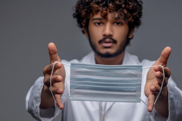 Profissional de saúde sério demonstrando, com um jaleco, uma máscara protetora descartável diante da câmera