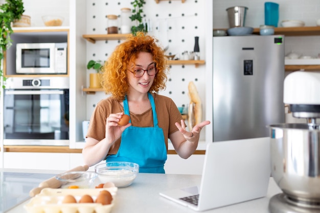 Profissional linda jovem feliz está blogando para seu canal de cozinha sobre vida saudável na cozinha de sua casa e olhando para a câmera em um laptop