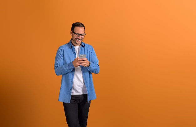 Profissional masculino alegre com óculos enviando mensagens por telefone inteligente e de pé em fundo laranja