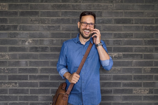 Profissional masculino feliz falando no telefone inteligente em pé contra a parede de tijolos