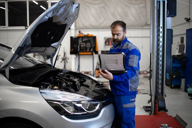 Profissional mecânico de automóveis caucasiano de meia idade com ferramenta de diagnóstico de computador portátil em pé pela área do motor do veículo com capô aberto, detectando mau funcionamento.