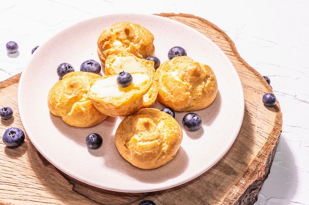 Profiteroles con arándanos. Pasteles de natillas, Eclair, hojaldre de crema sobre un soporte de madera, luz dura de moda, sombra oscura. Fondo de yeso blanco, cerrar