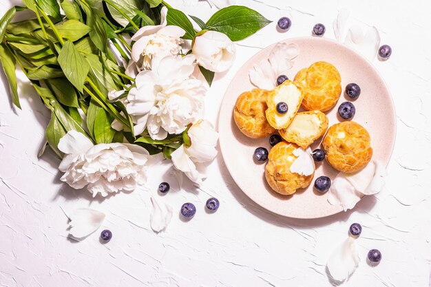 Profiteroles com mirtilos. Bolos de creme, Eclair, folhado de creme. Luz dura moderna, sombra escura. Peônias brancas em fundo de gesso, vista superior