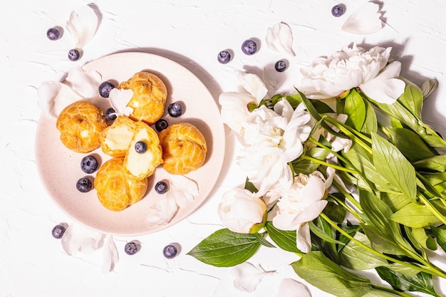 Profiteroles mit Blaubeeren. Vanillepuddingkuchen, Eclair, Windbeutel. Modernes hartes Licht, dunkler Schatten. Weiße Pfingstrosen auf Gipshintergrund, Ansicht von oben