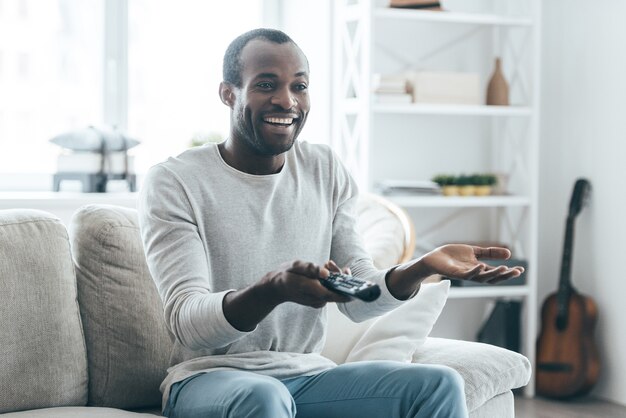 Programa de TV engraçado. Africano jovem bonito assistindo TV e sorrindo enquanto está sentado no sofá em casa