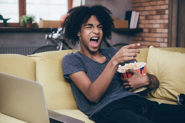 Programa de tv hilário. africano jovem alegre assistindo tv e segurando um balde de pipoca enquanto gesticula no sofá de casa