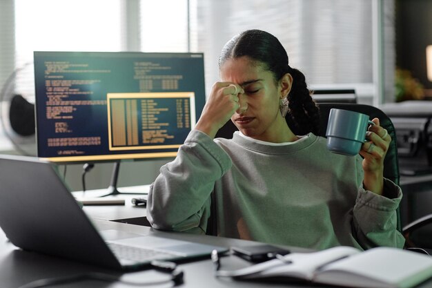 Programación femenina agotada tocando la cara sentada en el escritorio de trabajo en la oficina