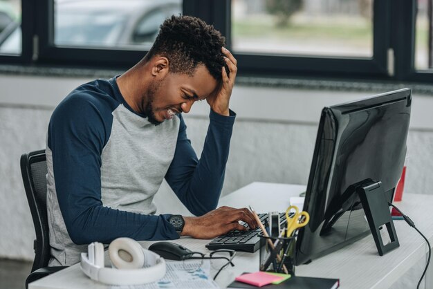 Programador afro-americano chateado segurando a mão perto da cabeça enquanto trabalhava no computador no escritório