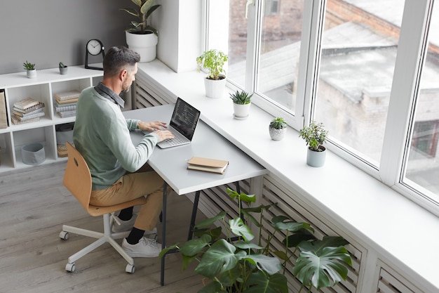 Programador de computador sentado em seu local de trabalho digitando em um laptop em um escritório espaçoso