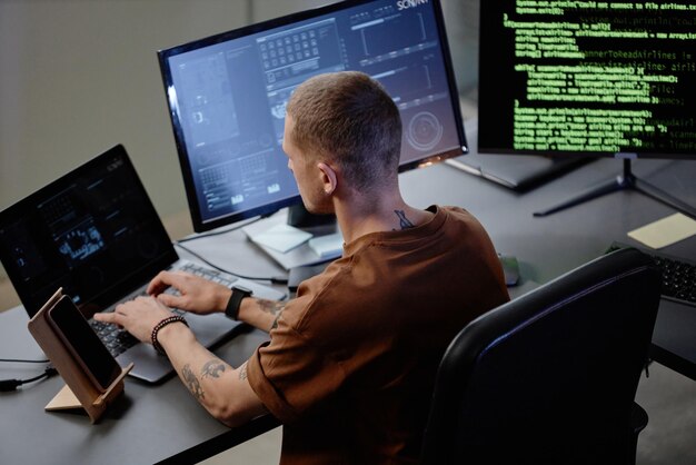 Programador de computadores masculino usando laptop em estação de trabalho com vários computadores no escritório