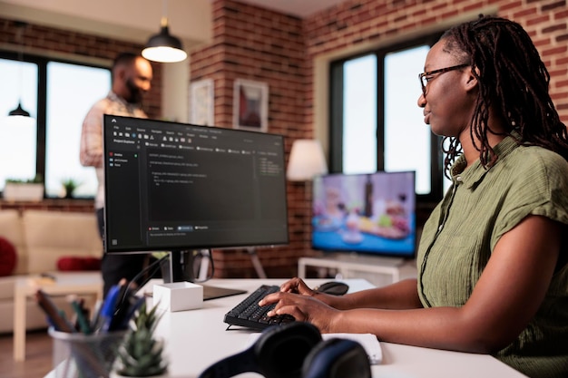 Programador de mulher trabalhando código de escrita remota usando computador enquanto namorado está relaxando assistindo tv. freelancer americano africano desenvolvendo software no pc sentado na mesa na sala de estar em casa