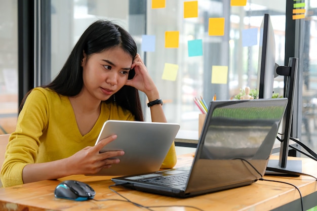 Programador feminino asiático usa camisas amarelas, olha para a tela do laptop e segura um tablet. ela mostrou uma maneira séria.