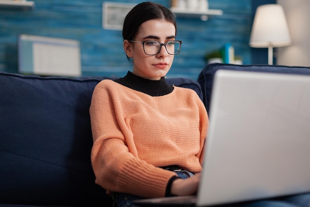 Programador con gafas sosteniendo portátil en sofá escribiendo código. Creador de contenido escribiendo en la oficina de la sala de estar de casa. Estudiante en el sofá mirando la publicación de la pantalla en las redes sociales.