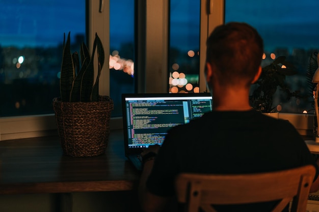 Foto un programador joven que codifica en una computadora portátil en la oscuridad con una vista de las luces de la ciudad de noche