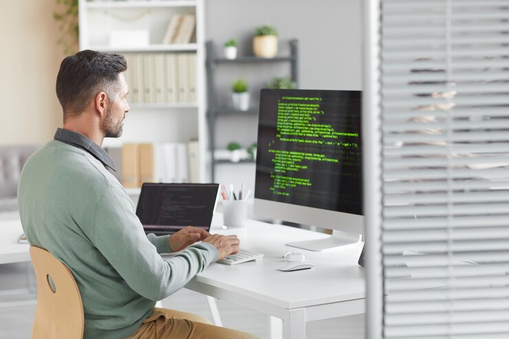 Foto programador joven sentado a la mesa frente al monitor de la computadora y desarrollando un nuevo sistema en la oficina