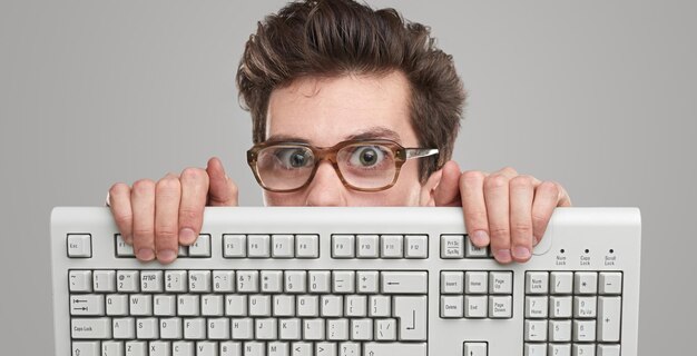 Programador masculino joven loco en gafas nerd mirando a la cámara desde detrás del teclado de la computadora contra el fondo gris