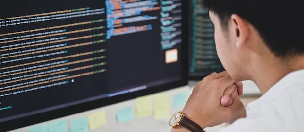 Un programador masculino se sienta y mira el código en un monitor de computadora para verificar la ejecución del programa.