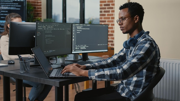 El programador piensa mientras toca la barba y arregla los anteojos mientras escribe en una computadora portátil sentada en el escritorio con múltiples pantallas de código de análisis. Administrador de base de datos enfocado que trabaja con la codificación del equipo en segundo plano.