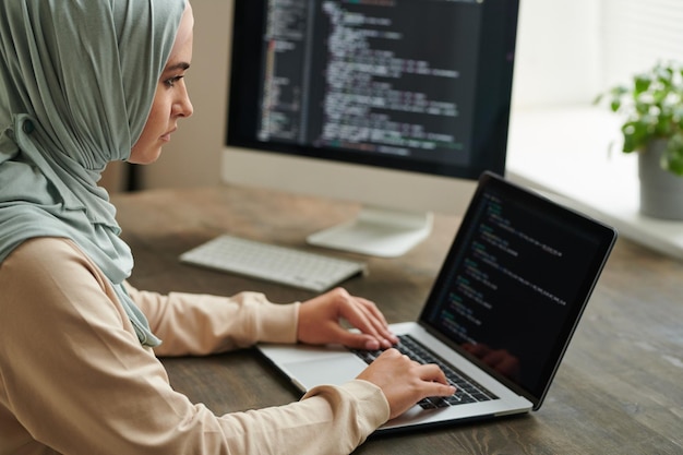 Programador profesional usando hiyab sentado en la mesa de la oficina escribiendo código de computadora en el espacio de copia de la computadora portátil