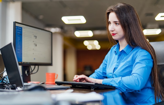 Programadora exitosa está sentada en el escritorio con una computadora mientras trabaja. Hermosa mujer mirando el monitor en la oficina de una empresa de software.