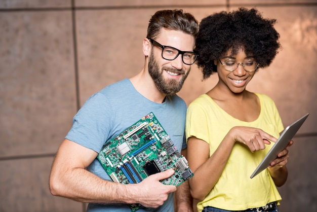 Foto programadores multiétnicos vestidos casualmente de pie junto con la placa base de la computadora y la tableta digital en el fondo de la pared en el interior