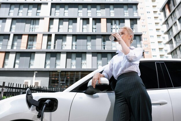 Progressive Frau mit Kaffee beim Aufladen von EV-Autos mit Wohngebäuden