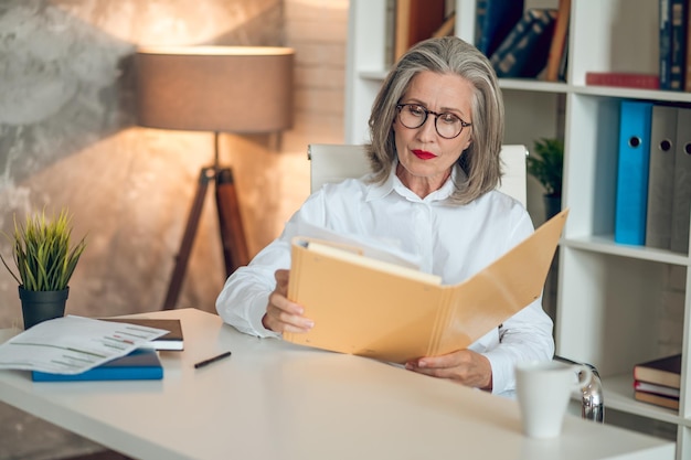 Projektmanager. Grauhaarige Frau, die am Tisch sitzt und mit der Projektdokumentation arbeitet