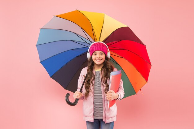 Projetado para clima chuvoso. Menina da escola segurar guarda-chuva colorido sobre fundo rosa. Criança pequena de volta à escola no outono. Criança adorável com fones de ouvido vai para a escola em dia chuvoso. A melhor escola.