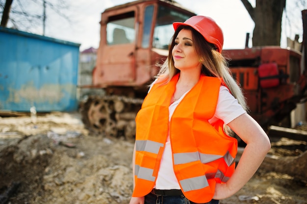 Projete a mulher do construtor no colete uniforme e no capacete protetor alaranjado contra a escavadora.