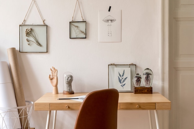 Projete o interior escandinavo de um escritório doméstico com várias molduras para fotos, mesa de madeira, cadeira marrom, plantas, escritório e acessórios pessoais. Encenação em casa neutra e elegante.