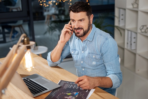 Projeto de negócios. Bonito e simpático empresário inteligente colocando um telefone no ouvido e falando com seu parceiro de negócios enquanto discutia o trabalho