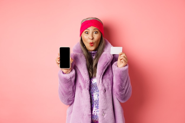 Promoción especial. Mujer mayor asiática emocionada que muestra la pantalla en blanco del teléfono inteligente y la tarjeta de crédito plástica, revisando la oferta en línea, fondo rosa
