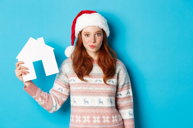 Promoções de férias e conceito imobiliário. Mulher ruiva bonita com chapéu de Papai Noel e camisola mostrando o modelo da casa de papel, oferta de apartamento, em pé sobre um fundo azul.